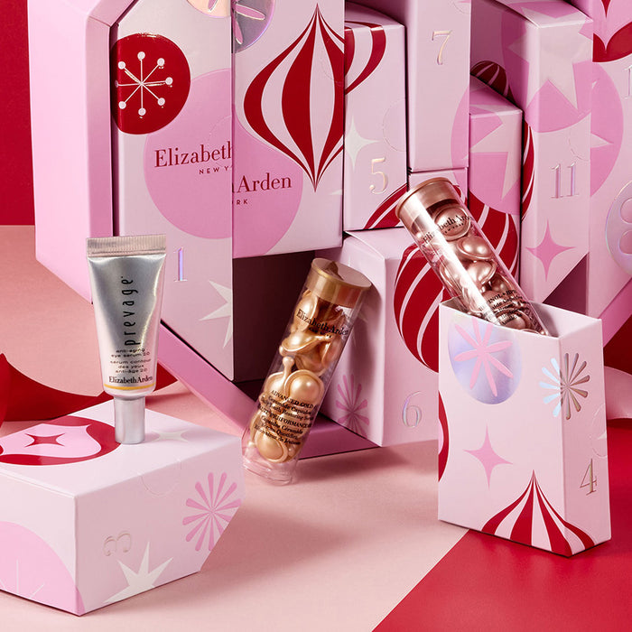 A zoomed in crop of the pink advent calendar gift box with multiple compartments, shown open with assorted cosmetic products inside. The box and surrounding props are decorated with pink, red, and white holiday-style graphics, set against a red and pink studio background.