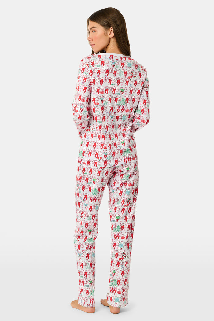 A woman stands outside with her arms crossed, wearing the Roller Rabbit Alpha Chi Omega Monkey Pajamas—a white Henley top and elastic waist pajama set featuring playful red and pink monkey patterns. She holds a matching quilted bag and carries a small heart-shaped blue and white polka dot purse.
