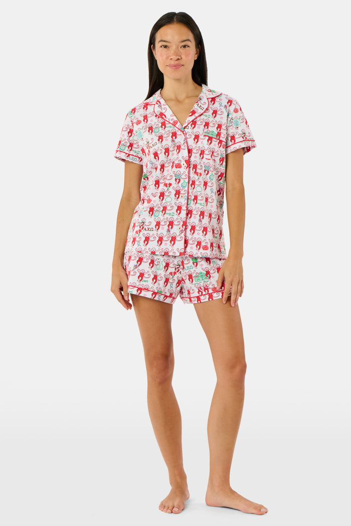 Wearing Roller Rabbit’s Alpha Chi Omega Monkey Polo Pajamas in a playful print, a woman leans against a brick wall, smiling softly as she holds two pink and white patterned notebooks and an iced coffee with a straw.