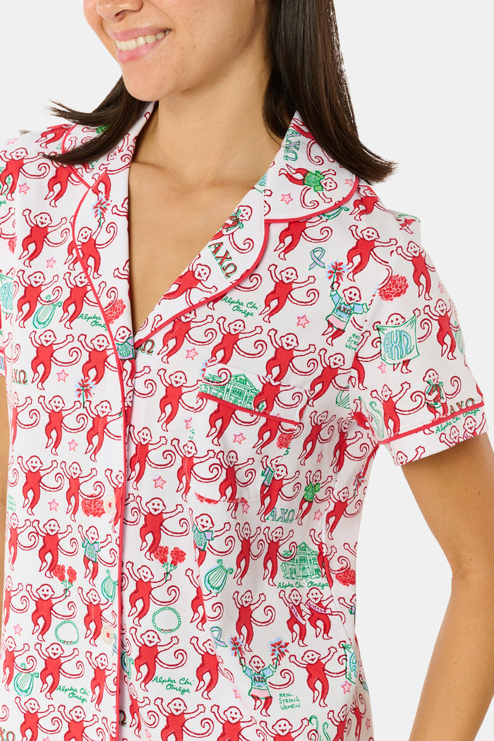 Wearing Roller Rabbit’s Alpha Chi Omega Monkey Polo Pajamas in a playful print, a woman leans against a brick wall, smiling softly as she holds two pink and white patterned notebooks and an iced coffee with a straw.