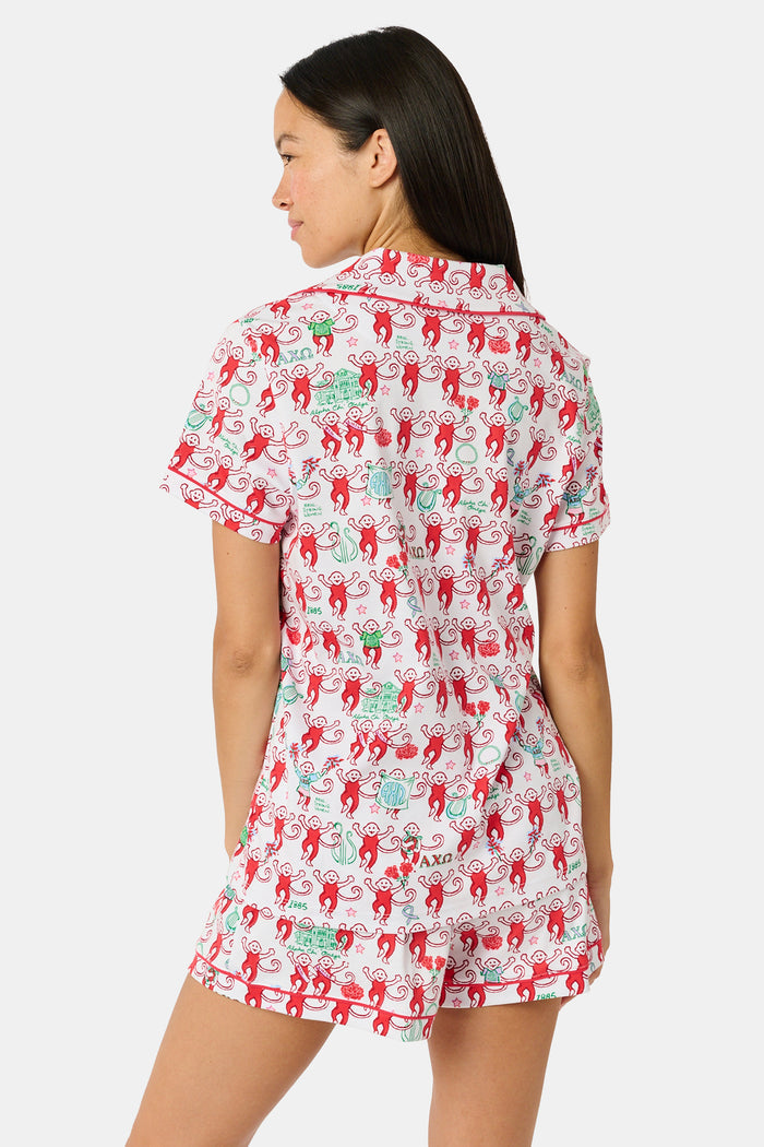Wearing Roller Rabbit’s Alpha Chi Omega Monkey Polo Pajamas in a playful print, a woman leans against a brick wall, smiling softly as she holds two pink and white patterned notebooks and an iced coffee with a straw.