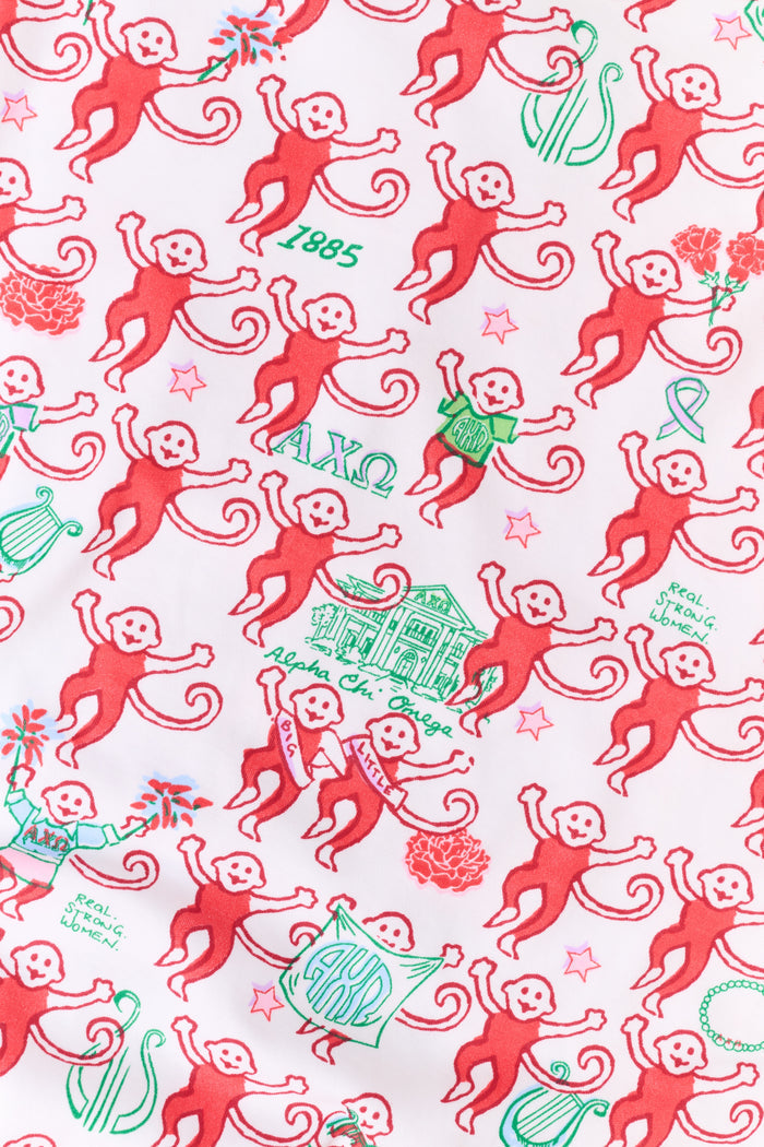 Wearing Roller Rabbit’s Alpha Chi Omega Monkey Polo Pajamas in a playful print, a woman leans against a brick wall, smiling softly as she holds two pink and white patterned notebooks and an iced coffee with a straw.