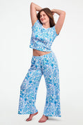 A woman stands barefoot on one leg, stretching with arms raised and eyes closed, wearing the Roller Rabbit Amanda Boden Pant and a matching blue floral short-sleeve pajama top.