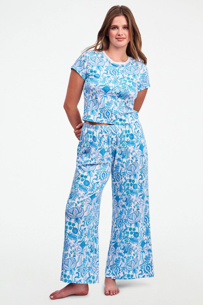 A woman stands barefoot on one leg, stretching with arms raised and eyes closed, wearing the Roller Rabbit Amanda Boden Pant and a matching blue floral short-sleeve pajama top.