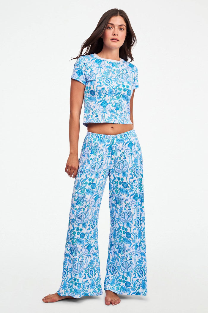 A woman stands barefoot on one leg, stretching with arms raised and eyes closed, wearing the Roller Rabbit Amanda Boden Pant and a matching blue floral short-sleeve pajama top.