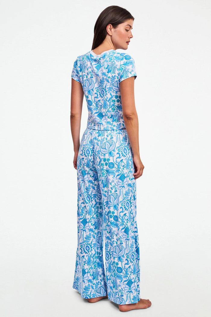 A woman stands barefoot on one leg, stretching with arms raised and eyes closed, wearing the Roller Rabbit Amanda Boden Pant and a matching blue floral short-sleeve pajama top.