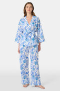 A woman poses barefoot against a plain white background wearing the Roller Rabbit Amanda Christy Set, a matching lounge set with a blue and white floral Amanda print, featuring a belted long-sleeve top and loose-fitting pants.