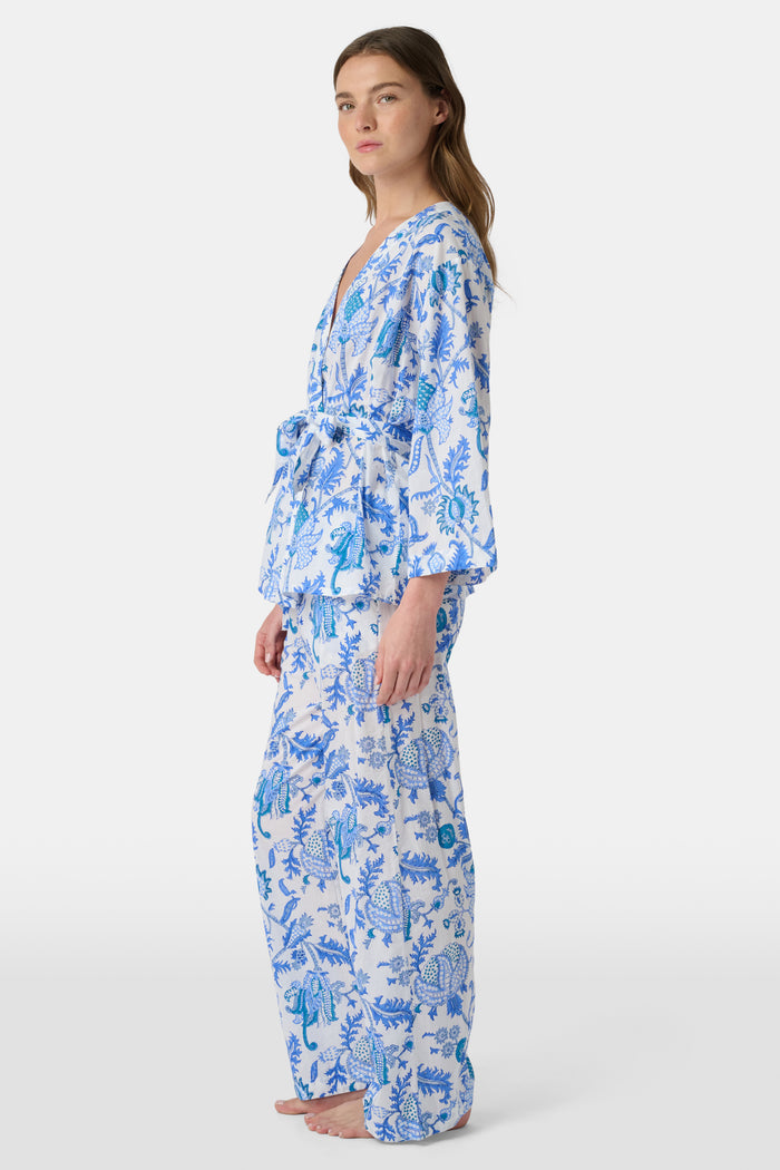 A woman poses barefoot against a plain white background wearing the Roller Rabbit Amanda Christy Set, a matching lounge set with a blue and white floral Amanda print, featuring a belted long-sleeve top and loose-fitting pants.