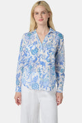 A woman with long blonde hair wears the Roller Rabbit Amanda Guy Shirt, a light blue and white paisley long-sleeve top, paired with white pants, standing against a plain white background.