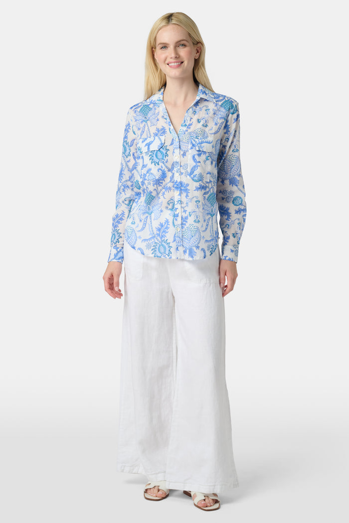 A woman with long blonde hair wears the Roller Rabbit Amanda Guy Shirt, a light blue and white paisley long-sleeve top, paired with white pants, standing against a plain white background.