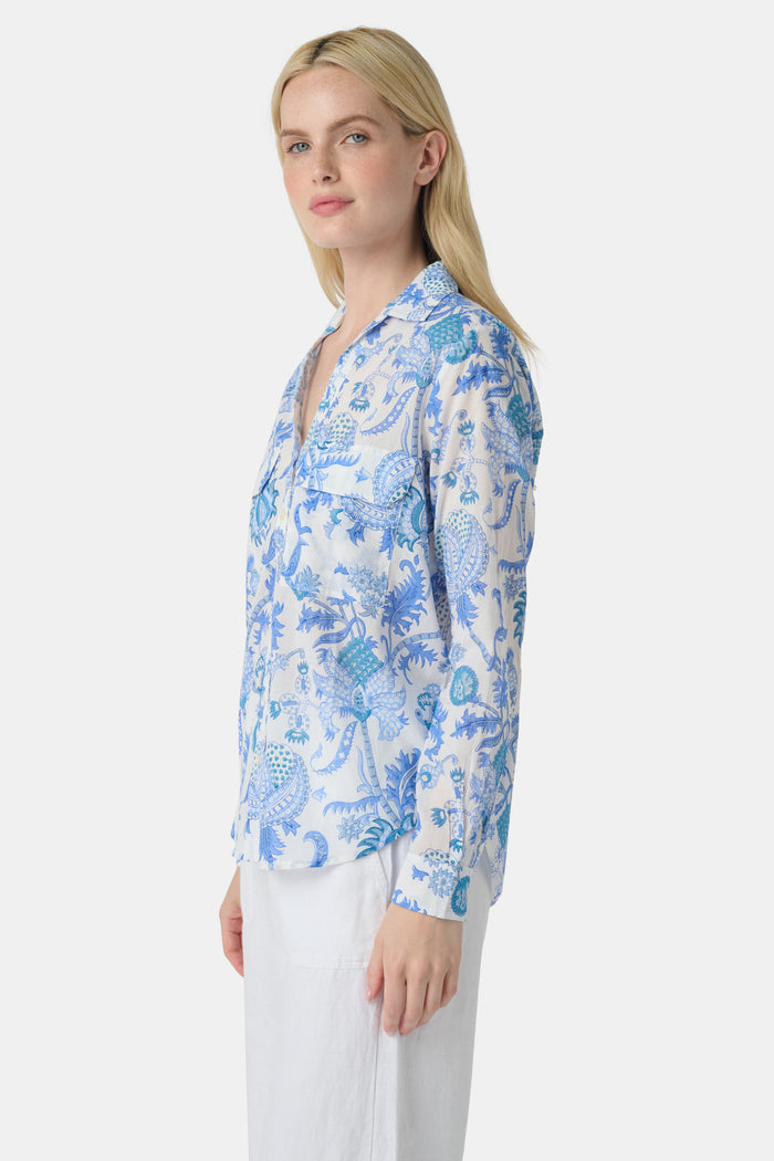 A woman with long blonde hair wears the Roller Rabbit Amanda Guy Shirt, a light blue and white paisley long-sleeve top, paired with white pants, standing against a plain white background.