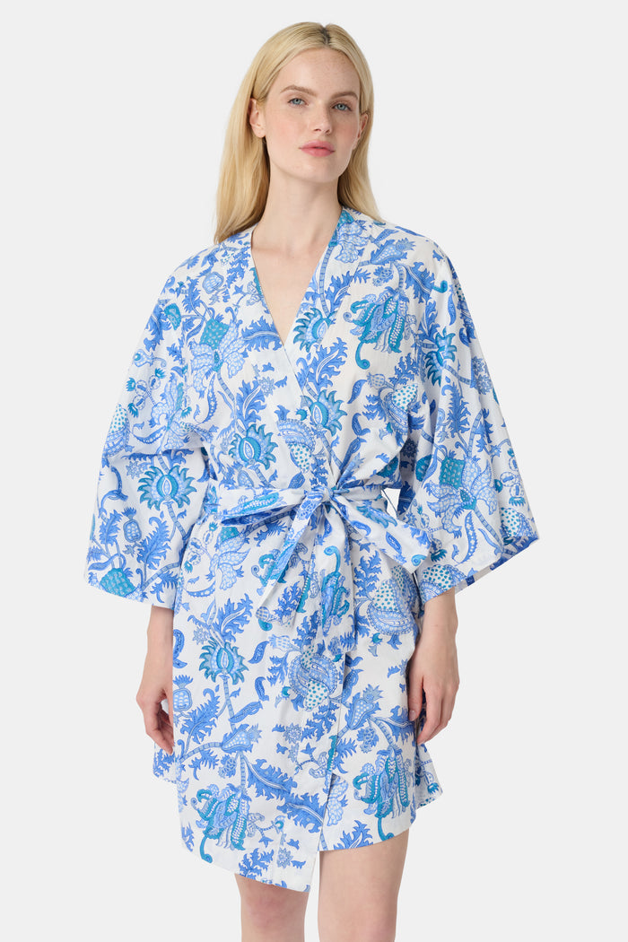 A woman stands barefoot against a white background, wearing the Roller Rabbit Amanda Kimono—a knee-length cotton loungewear piece with blue-and-white floral patterns, wide sleeves, and a belt.
