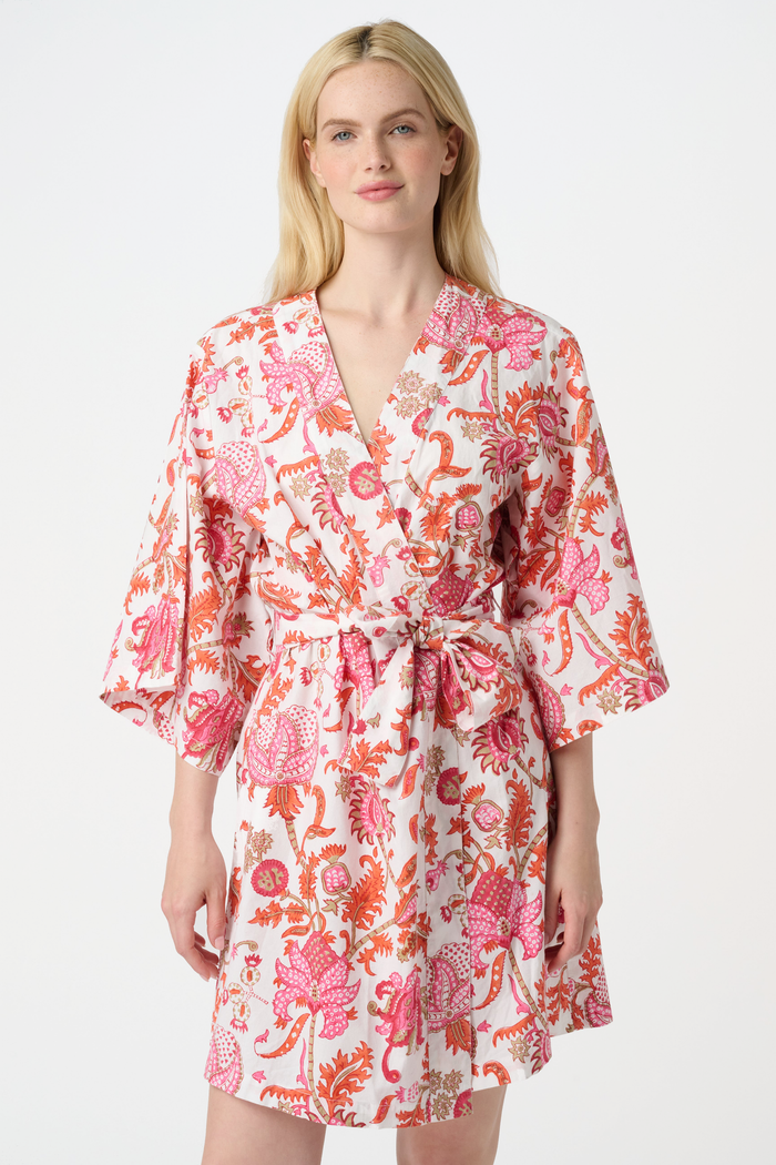 A woman with long blonde hair wears the Roller Rabbit Amanda Kimono, a belted, knee-length cotton kimono with a vibrant pink and orange floral pattern on white. She stands against a plain light gray backdrop, smiling softly.