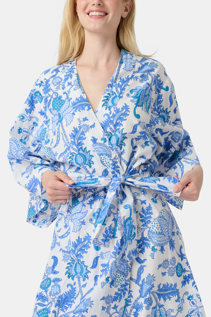 A woman stands barefoot against a white background, wearing the Roller Rabbit Amanda Kimono—a knee-length cotton loungewear piece with blue-and-white floral patterns, wide sleeves, and a belt.