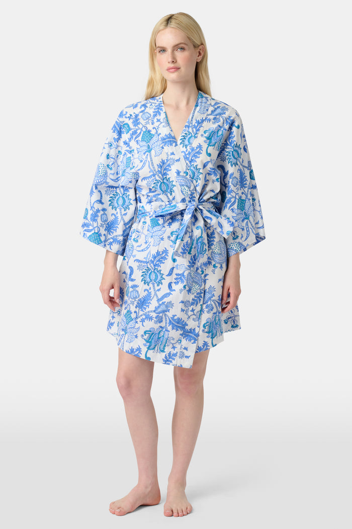 A woman stands barefoot against a white background, wearing the Roller Rabbit Amanda Kimono—a knee-length cotton loungewear piece with blue-and-white floral patterns, wide sleeves, and a belt.