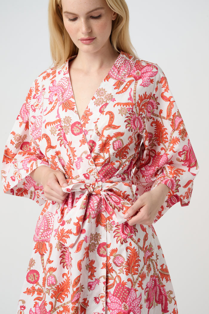 A woman with long blonde hair wears the Roller Rabbit Amanda Kimono, a belted, knee-length cotton kimono with a vibrant pink and orange floral pattern on white. She stands against a plain light gray backdrop, smiling softly.