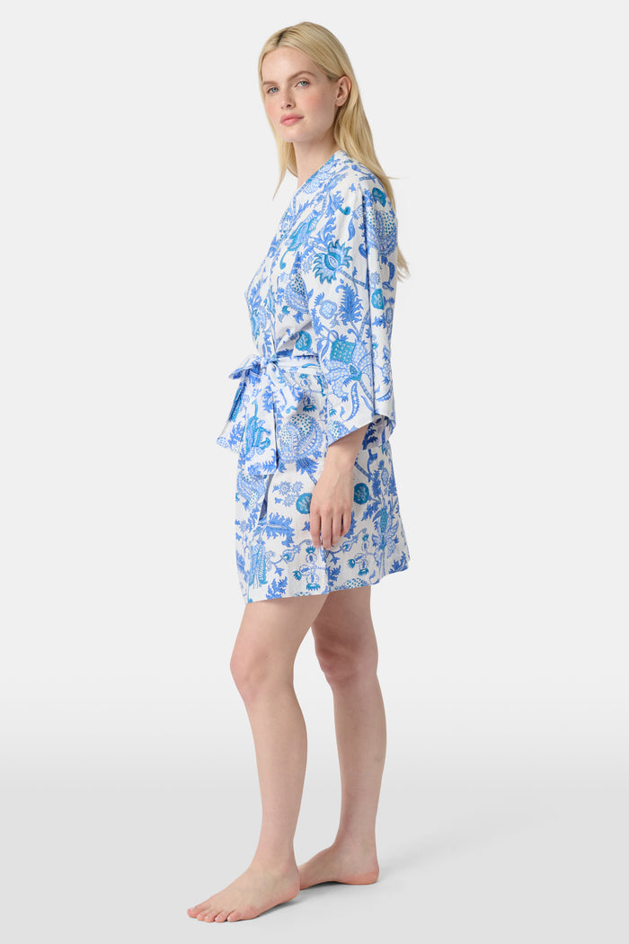 A woman stands barefoot against a white background, wearing the Roller Rabbit Amanda Kimono—a knee-length cotton loungewear piece with blue-and-white floral patterns, wide sleeves, and a belt.
