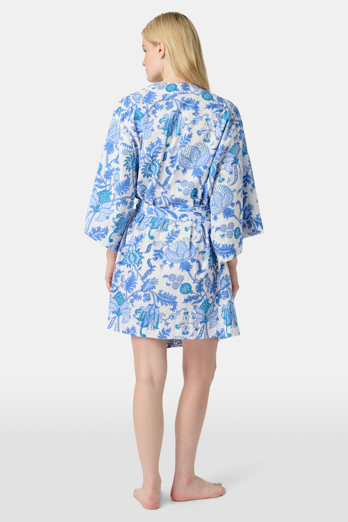 A woman stands barefoot against a white background, wearing the Roller Rabbit Amanda Kimono—a knee-length cotton loungewear piece with blue-and-white floral patterns, wide sleeves, and a belt.