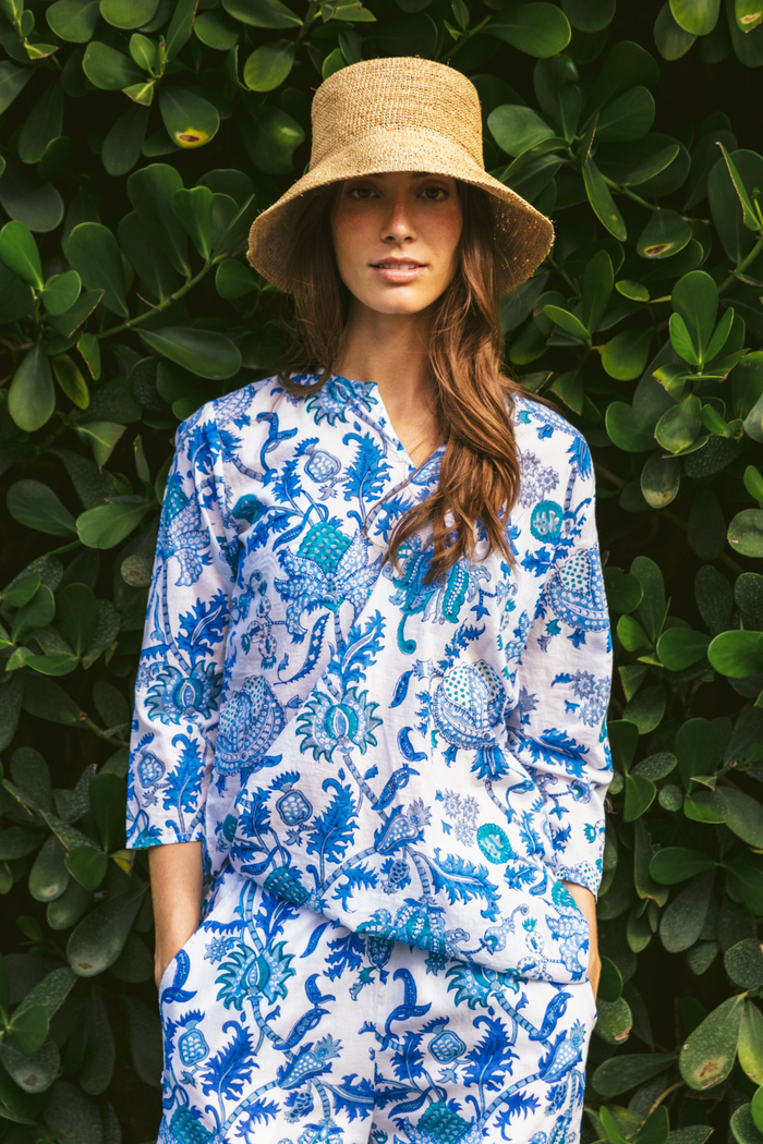 A woman poses in front of green leaves, wearing the Amanda Kurta by Roller Rabbit—a 100 cotton blue-and-white floral-patterned piece with hand-beaded accents—paired with a straw hat and hands in her pockets.