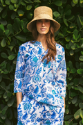A woman poses in front of green leaves, wearing the Amanda Kurta by Roller Rabbit—a 100 cotton blue-and-white floral-patterned piece with hand-beaded accents—paired with a straw hat and hands in her pockets.