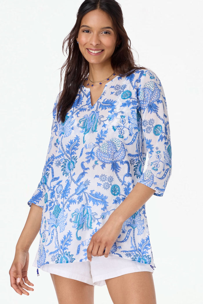 A woman poses in front of green leaves, wearing the Amanda Kurta by Roller Rabbit—a 100 cotton blue-and-white floral-patterned piece with hand-beaded accents—paired with a straw hat and hands in her pockets.