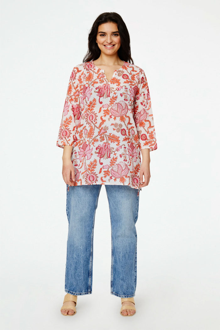 A woman with long dark hair smiles, seated on a red and pink patterned blanket. She wears the Roller Rabbit Amanda Kurta—a light pink 100 cotton top with red and orange floral print—white shorts, and sits against a plain white background.