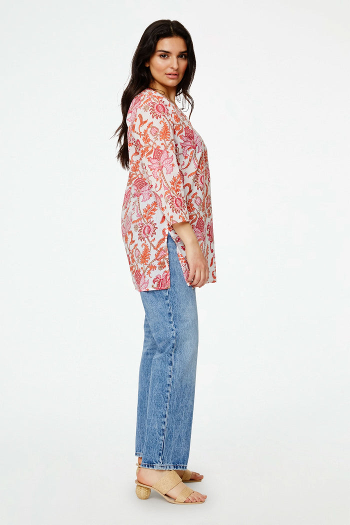 A woman with long dark hair smiles, seated on a red and pink patterned blanket. She wears the Roller Rabbit Amanda Kurta—a light pink 100 cotton top with red and orange floral print—white shorts, and sits against a plain white background.