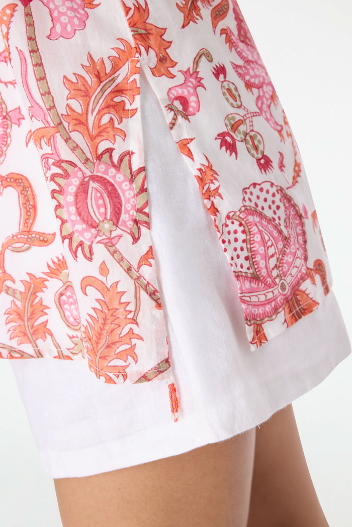 A woman with long dark hair smiles, seated on a red and pink patterned blanket. She wears the Roller Rabbit Amanda Kurta—a light pink 100 cotton top with red and orange floral print—white shorts, and sits against a plain white background.