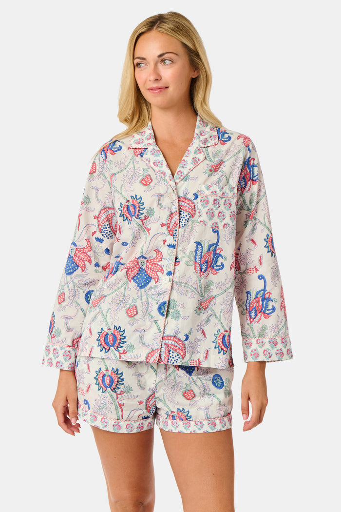 A woman stands against a plain white background, smiling slightly as she models the Amanda Leila Set from Roller Rabbit—a vintage-inspired cotton loungewear ensemble featuring a long-sleeve, button-up pajama top and matching shorts adorned with a colorful floral pattern.