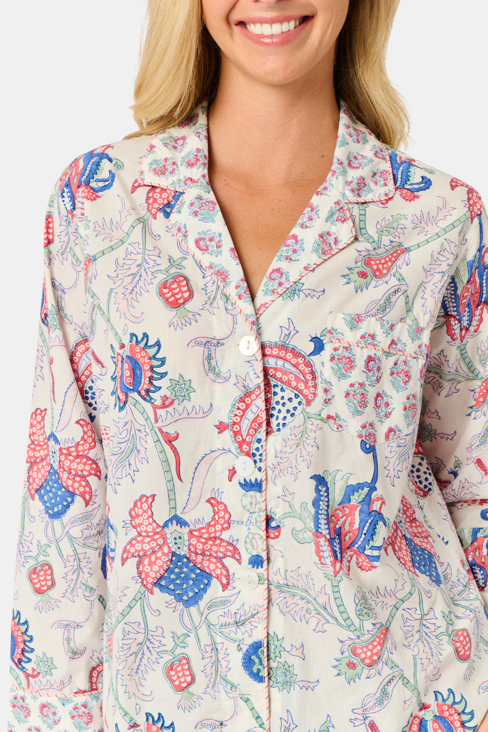 A woman stands against a plain white background, smiling slightly as she models the Amanda Leila Set from Roller Rabbit—a vintage-inspired cotton loungewear ensemble featuring a long-sleeve, button-up pajama top and matching shorts adorned with a colorful floral pattern.
