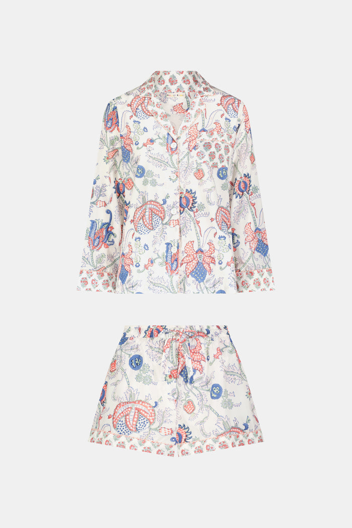A woman stands against a plain white background, smiling slightly as she models the Amanda Leila Set from Roller Rabbit—a vintage-inspired cotton loungewear ensemble featuring a long-sleeve, button-up pajama top and matching shorts adorned with a colorful floral pattern.