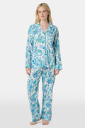 A woman with blonde hair and a neutral expression wears the Roller Rabbit Amanda Loungewear—a white cotton pajama set with a blue and green botanical print, featuring a button-up long-sleeve top and matching pants. She stands barefoot.