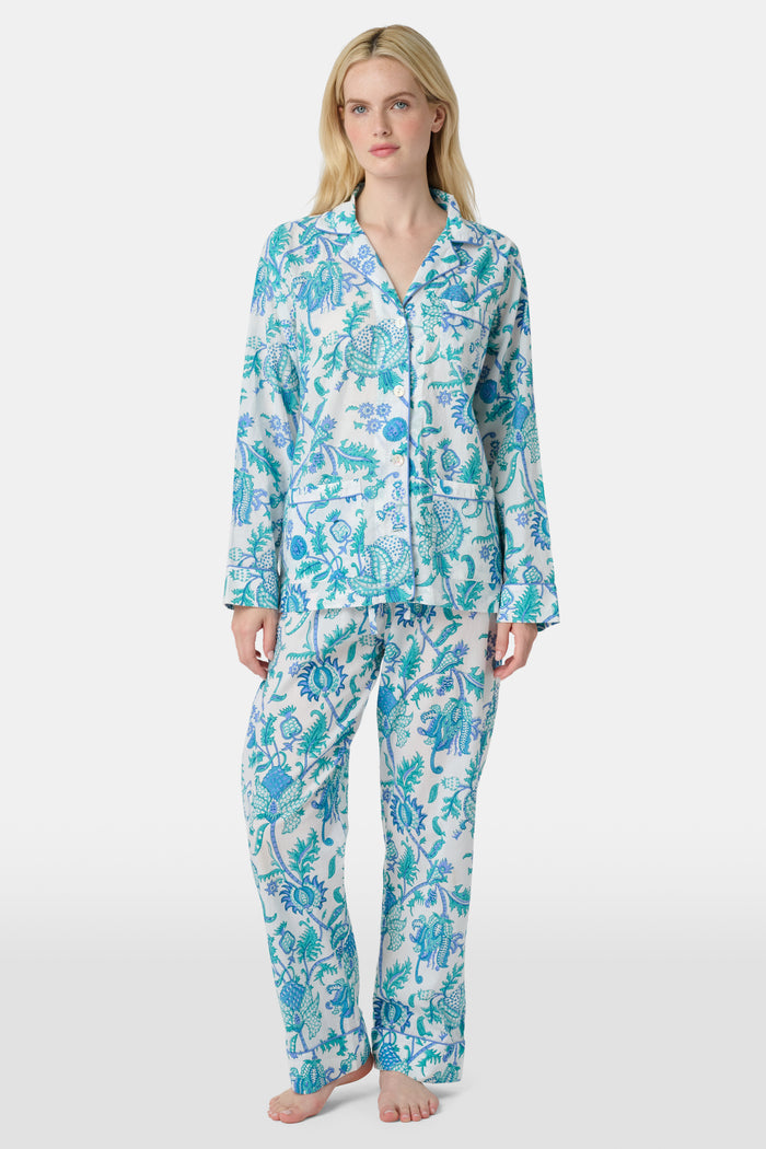 A woman with blonde hair and a neutral expression wears the Roller Rabbit Amanda Loungewear—a white cotton pajama set with a blue and green botanical print, featuring a button-up long-sleeve top and matching pants. She stands barefoot.