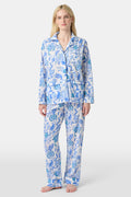 A woman stands barefoot in the Amanda Loungewear set by Roller Rabbit, featuring a blue and white botanical print, long-sleeve collared button-up shirt, and matching pants against a plain light background.