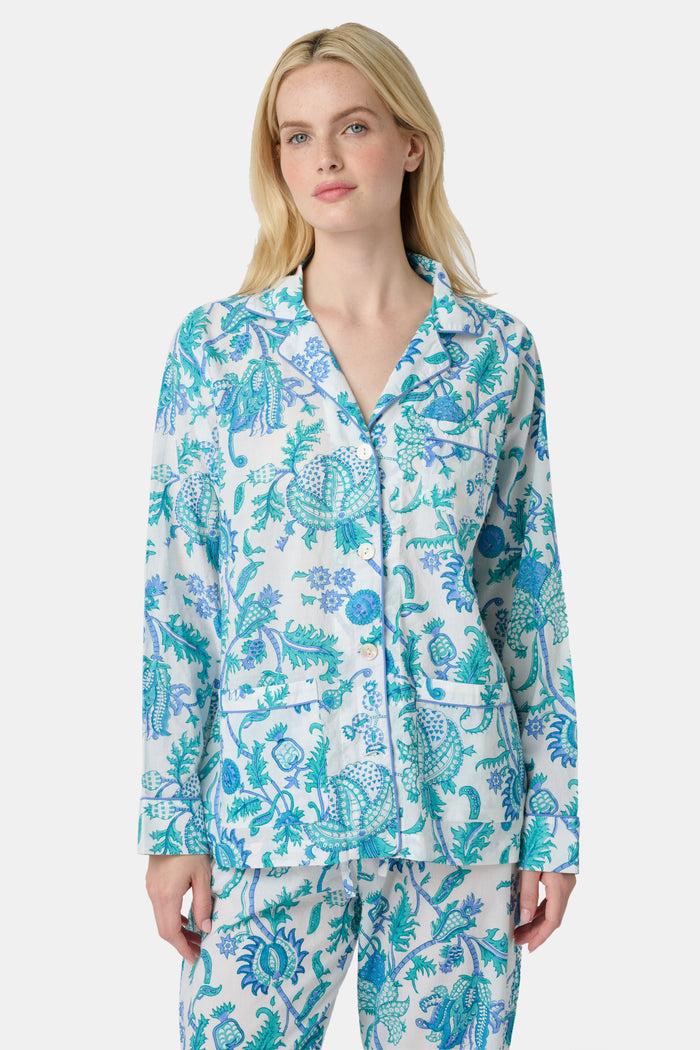 A woman with blonde hair and a neutral expression wears the Roller Rabbit Amanda Loungewear—a white cotton pajama set with a blue and green botanical print, featuring a button-up long-sleeve top and matching pants. She stands barefoot.