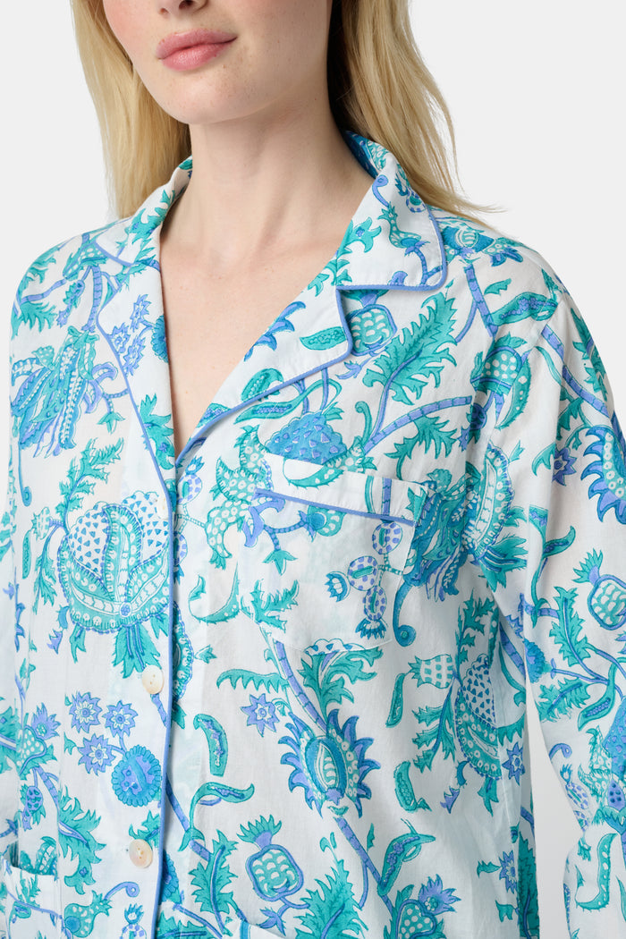 A woman with blonde hair and a neutral expression wears the Roller Rabbit Amanda Loungewear—a white cotton pajama set with a blue and green botanical print, featuring a button-up long-sleeve top and matching pants. She stands barefoot.