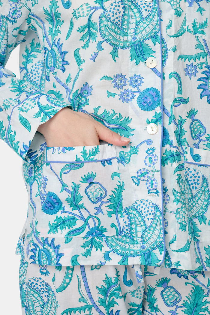 A woman with blonde hair and a neutral expression wears the Roller Rabbit Amanda Loungewear—a white cotton pajama set with a blue and green botanical print, featuring a button-up long-sleeve top and matching pants. She stands barefoot.