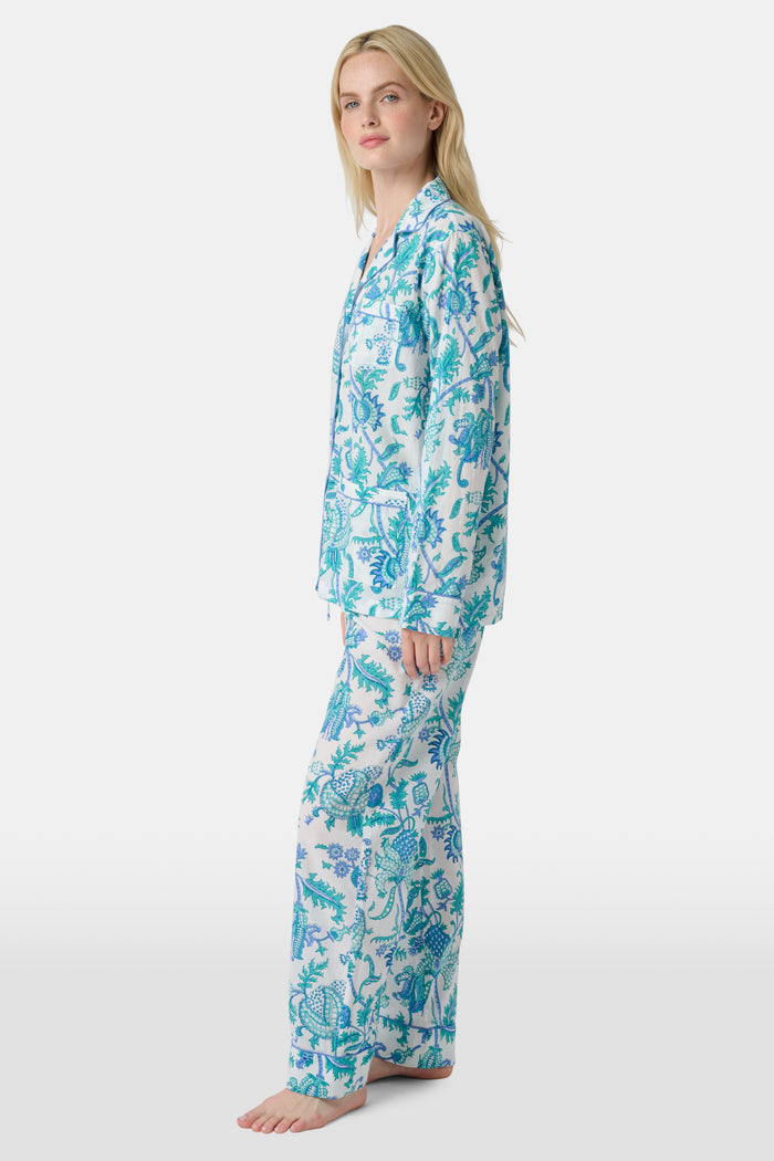 A woman with blonde hair and a neutral expression wears the Roller Rabbit Amanda Loungewear—a white cotton pajama set with a blue and green botanical print, featuring a button-up long-sleeve top and matching pants. She stands barefoot.