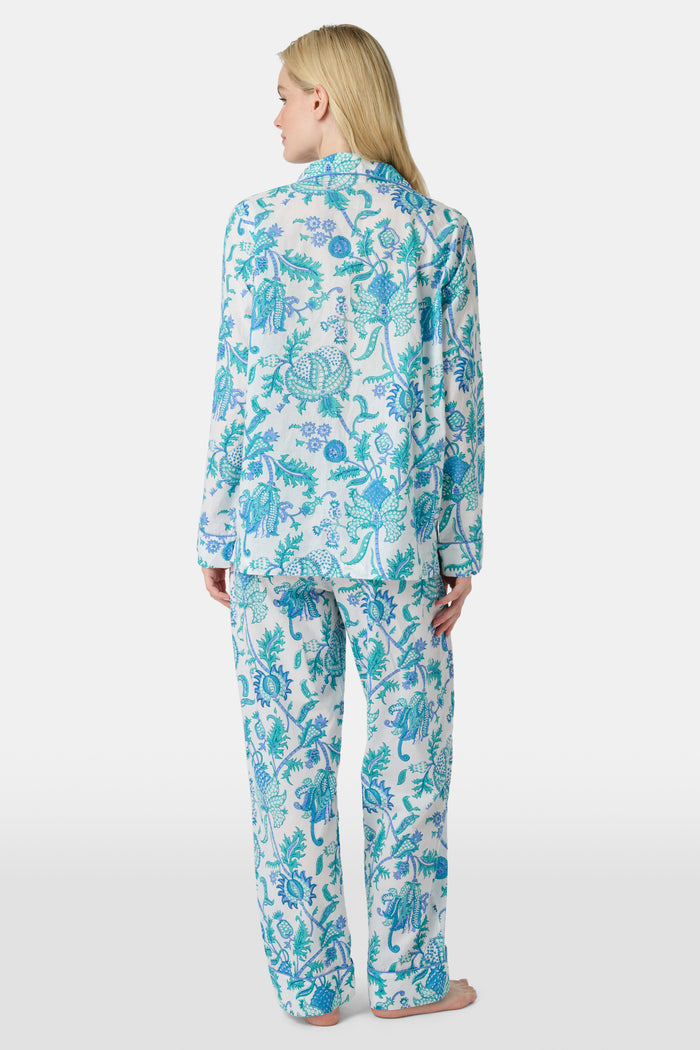 A woman with blonde hair and a neutral expression wears the Roller Rabbit Amanda Loungewear—a white cotton pajama set with a blue and green botanical print, featuring a button-up long-sleeve top and matching pants. She stands barefoot.