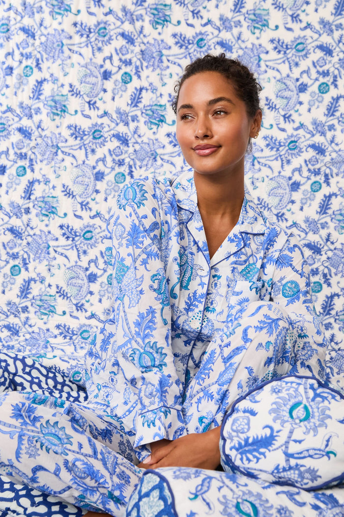 A woman stands barefoot in the Amanda Loungewear set by Roller Rabbit, featuring a blue and white botanical print, long-sleeve collared button-up shirt, and matching pants against a plain light background.