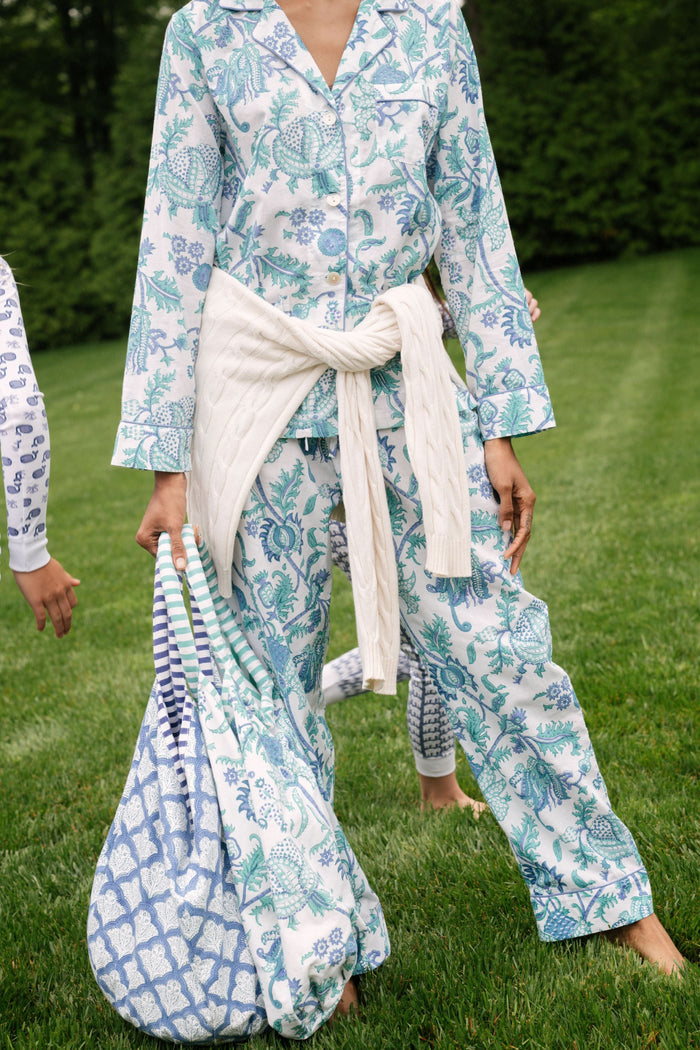 A woman with blonde hair and a neutral expression wears the Roller Rabbit Amanda Loungewear—a white cotton pajama set with a blue and green botanical print, featuring a button-up long-sleeve top and matching pants. She stands barefoot.