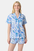 A woman with long blonde hair smiles in the Amanda Lulu Set by Roller Rabbit, a blue floral print cotton short set, standing against a plain light background and showcasing this chic women's loungewear.