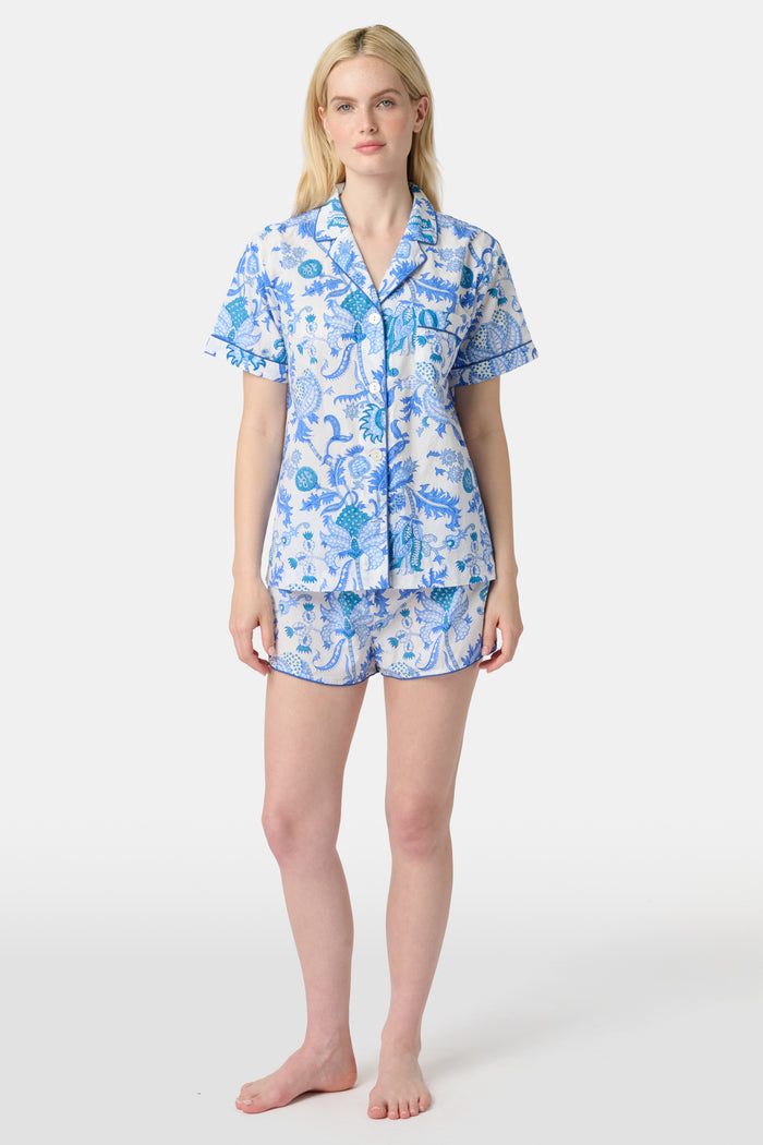 A woman with long blonde hair smiles in the Amanda Lulu Set by Roller Rabbit, a blue floral print cotton short set, standing against a plain light background and showcasing this chic women's loungewear.