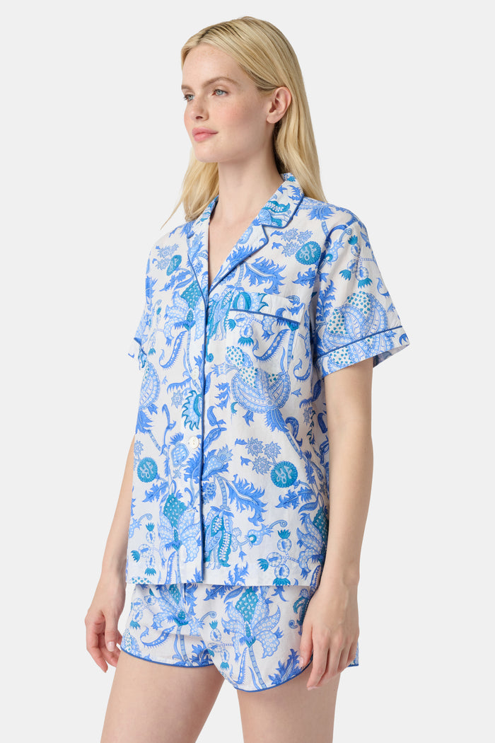 A woman with long blonde hair smiles in the Amanda Lulu Set by Roller Rabbit, a blue floral print cotton short set, standing against a plain light background and showcasing this chic women's loungewear.