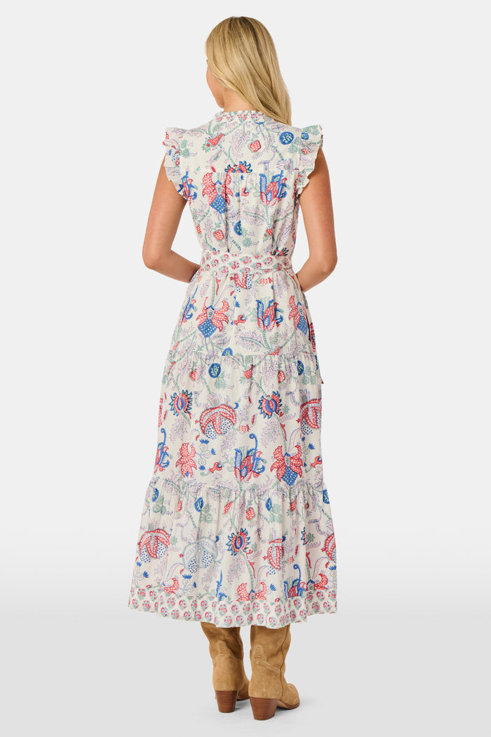 A person in the Amanda Nolan Dress by Roller Rabbit—a patterned, sleeveless, light-colored cotton maxi—stands on grass, holding out one side of the skirt. A wooden fence and lush greenery form the background; their face is not visible.