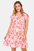 A woman wears the Roller Rabbit Amanda Pamela Dress—a short-sleeved, knee-length, tiered cotton summer dress with a pink and red floral print—standing against a plain light background, smiling with one hand on the hem.