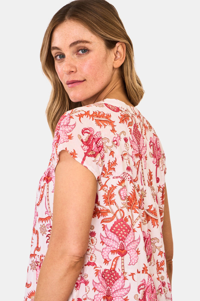 A woman wears the Roller Rabbit Amanda Pamela Dress—a short-sleeved, knee-length, tiered cotton summer dress with a pink and red floral print—standing against a plain light background, smiling with one hand on the hem.
