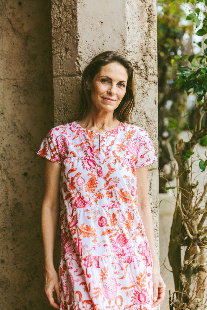 A woman wears the Roller Rabbit Amanda Pamela Dress—a short-sleeved, knee-length, tiered cotton summer dress with a pink and red floral print—standing against a plain light background, smiling with one hand on the hem.