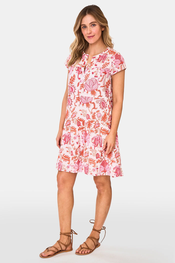 A woman wears the Roller Rabbit Amanda Pamela Dress—a short-sleeved, knee-length, tiered cotton summer dress with a pink and red floral print—standing against a plain light background, smiling with one hand on the hem.