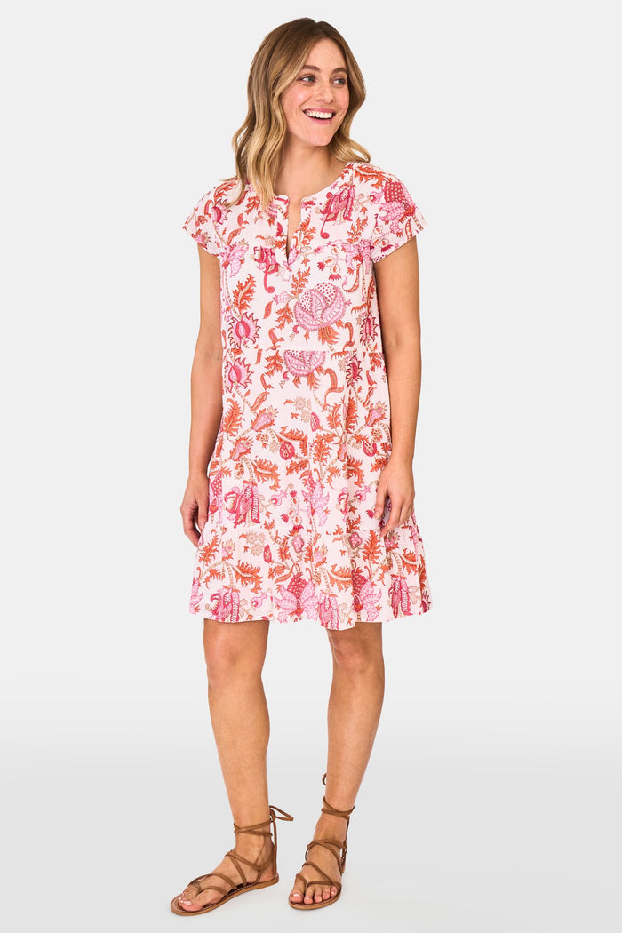 A woman wears the Roller Rabbit Amanda Pamela Dress—a short-sleeved, knee-length, tiered cotton summer dress with a pink and red floral print—standing against a plain light background, smiling with one hand on the hem.